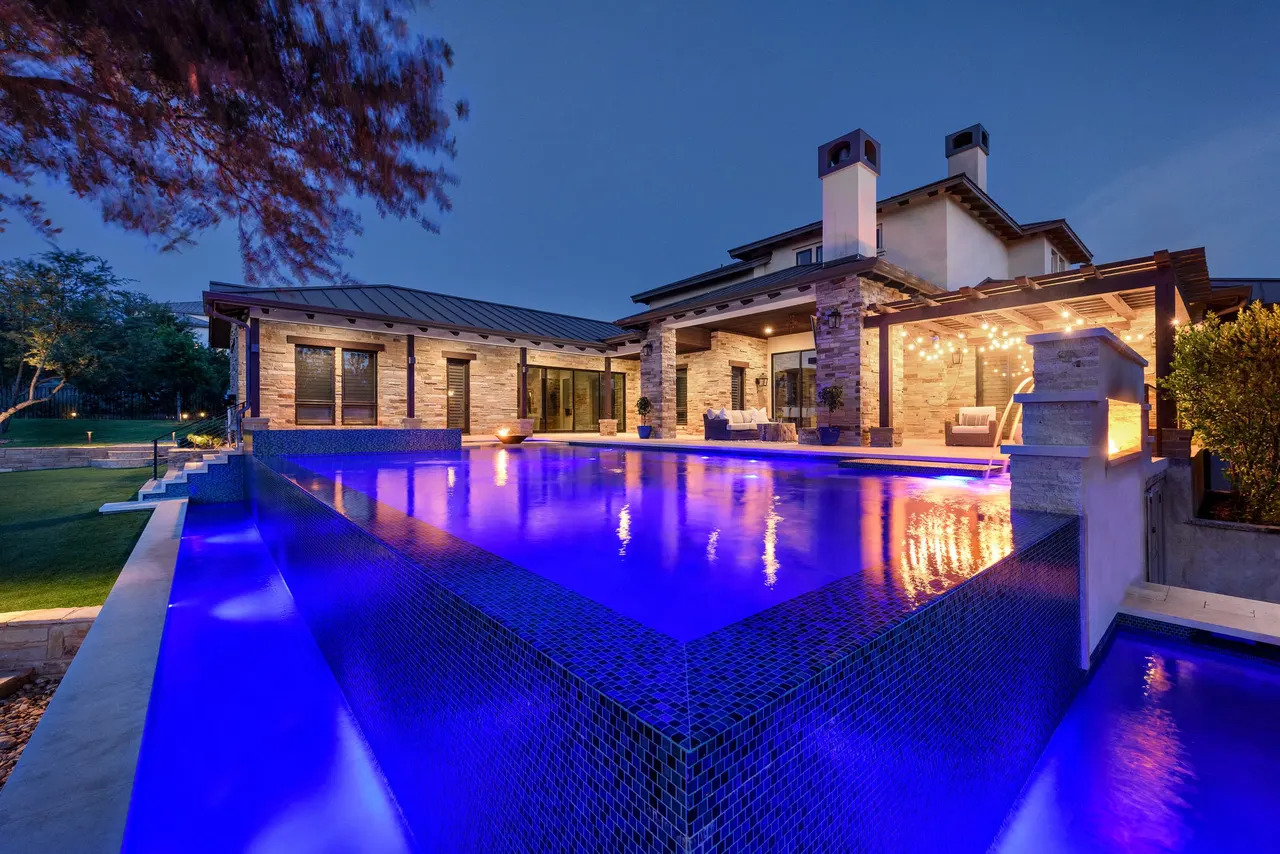Infinity-edge estate pool lit at dusk with fire bowls on the weir and a stone water wall — Fire & Water Estate, Texas Hill Country